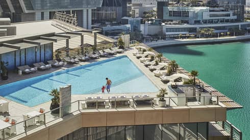 A wide shot of a man and child walking around a triangular pool overlooking the water and Abu Dhabi skyscrapers in the background. 