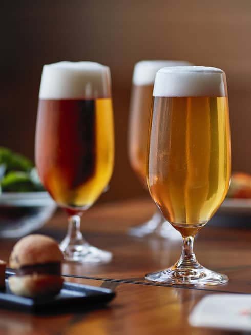 Four tall beer glasses filled with beer on a table with cheese, crackers and appetizers