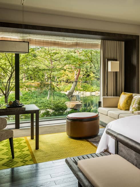 Guest room with a king bed, sofa with green cushions, round ottoman and large window overlooking a pond garden