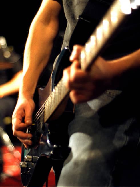 Close-up of musician's arms and hands playing an electric guitar with drumset in the background.