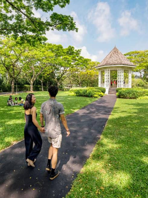 A man and woman hold hands and walk along an asphalt trail through the Singapore Botanic Gardens