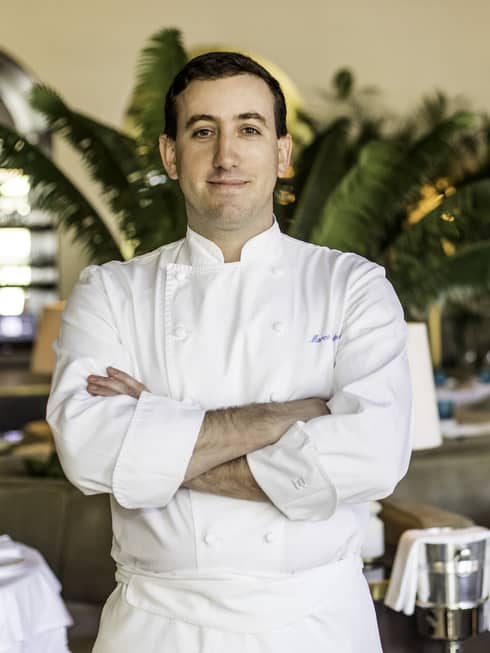 Chef Marco Calenzi stands in one of the Surfside restaurants at Four Seasons Surf Club. He is wearing a chef's coat and the restaurant has plants and white cloth tables. 