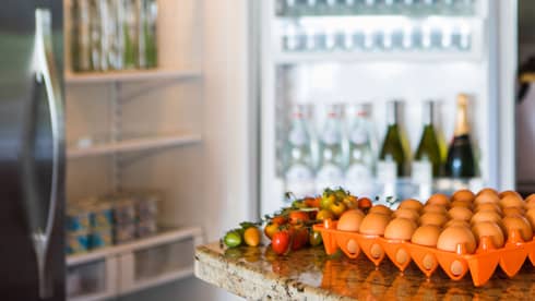 Smiling man holds green bottle in front of open fridge, fresh eggs on counter