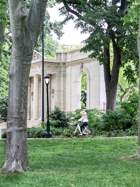 A person riding a bike through a park with trees and old stone architecture.