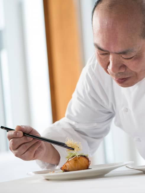 Chef in white uniform leans over grilled salmon dish on plate, adds garnish with chopsticks