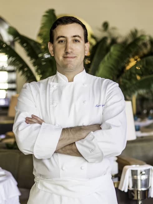 A chef in a chef's coat in a restaurant with plants and white cloth tables.
