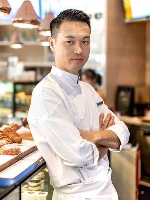 A male Asian chef standing with his arms crossed, his back to a tray of assorted pastries