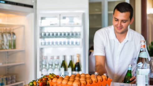 Smiling man holds green bottle in front of open fridge, fresh eggs on counter