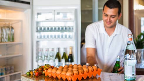 Smiling man holds green bottle in front of open fridge, fresh eggs on counter