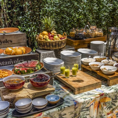 Outdoor table covered in a floral table cloth is filled with stacks of white dishes, wooden platters topped with bowls of food and an assortment of brunch items on display