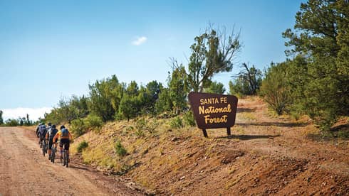Group of cyclists ride up dirt path in a row past Santa Fe National Forest sign 