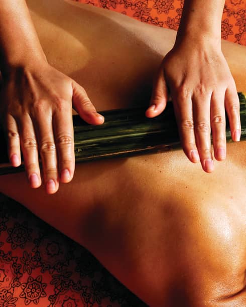 Spa staff rolls thick bamboo stick over woman's bare back, shoulders 