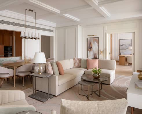 Elegant open-plan living space with neutral furnishings, pastel accents, a marble kitchen island and a dining area beside a modern chandelier.