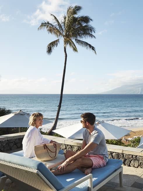 Man in shorts and smiling woman in beach shirt sit on edge of lounge chair, beach and ocean in background