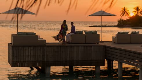 Private island dining at sunset. Woman with long dress blowing in the wind, man sits on the edge of white sofa