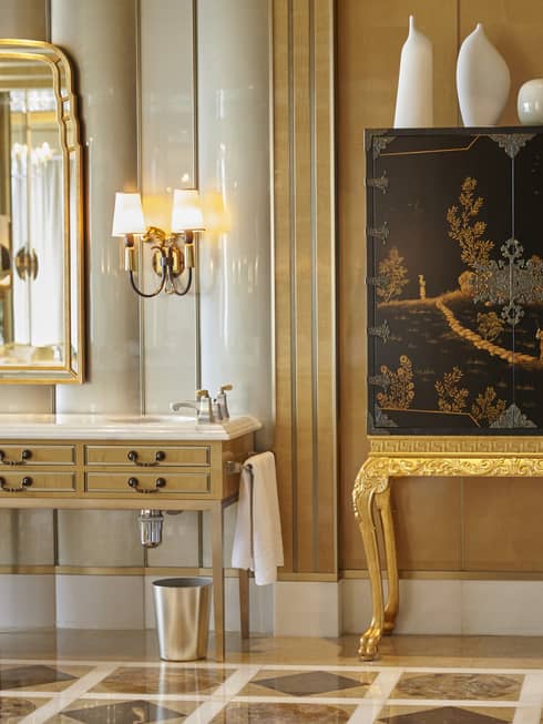 Elegant bathroom with patterned marble floors, large gold mirror over sink, chinoiserie cabinet topped with modern white vases