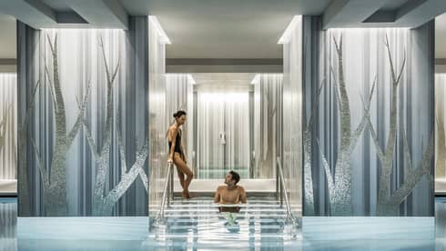 A couple enjoys a serene swimming pool, with one person seated on steps between elegant blue and silver columns.
