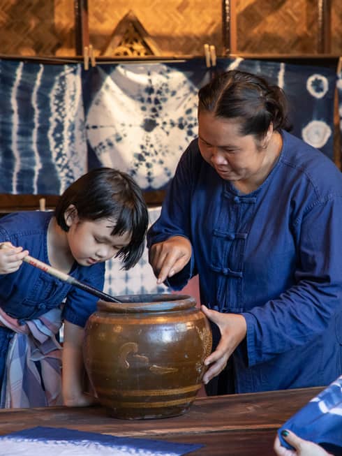 A woman teaching a child how to dye fabric using indigo, with tie-dyed cloths hanging in the background in a traditional workshop.