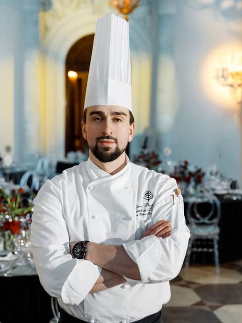 Chef Denis, dressed in a chefs hat and apron, stands in the middle of a restaurant with his arms crossed against his white chef's jacket..