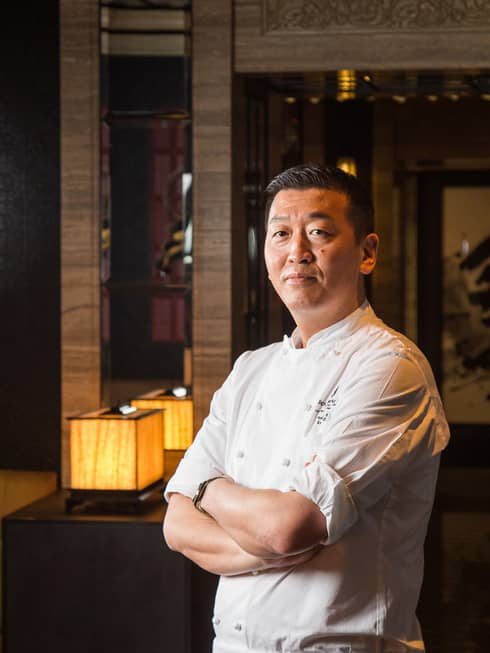 Chef Toshikazu Kato in chef's coat with arms folded, Asian restaurant interior in backdrop