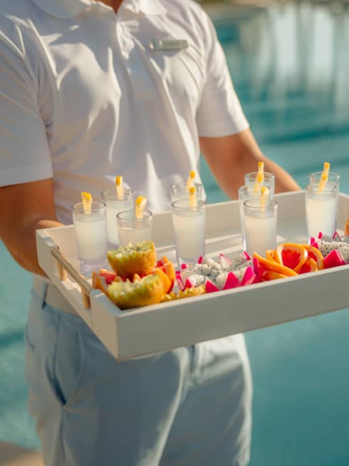 Pool attendant wearing a white short-sleeve polo shirt and light-blue shorts carries a large tray filled with beverages and small plates of colourful fruit and snacks
