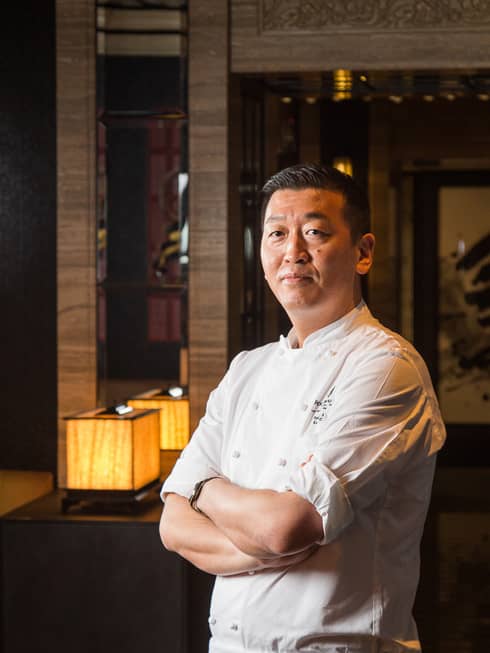 Chef Toshikazu Kato in chef's coat with arms folded, Asian restaurant interior in backdrop