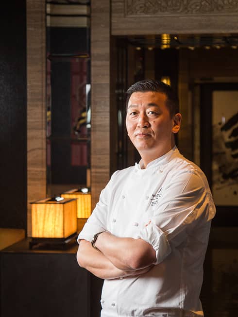 Chef Toshikazu Kato in chef's coat with arms folded, Asian restaurant interior in backdrop