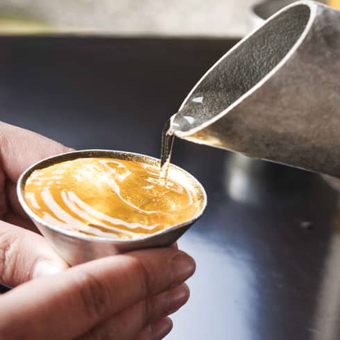 Clear liquid taking on amber colour as it is poured from a hammered metal spout into small cone-shaped cup held by two hands