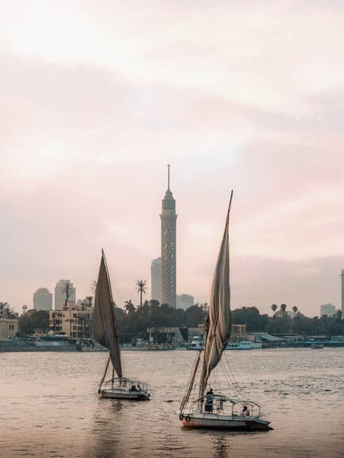 Two felucca boats drift on the water in the evening light, a busy shoreline and Cairo Tower in the distance. 