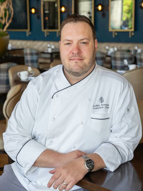 A male chef in a white chefs coat sitting at a counter of a restaurant.