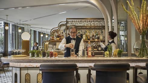 Two bartenders, one male and one female, prepare cocktails at Miss River's marble-topped bar, liquor bottles arranged behind them