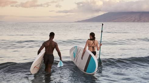 Two people standing in the ocean holding paddleboards with mountains in the background