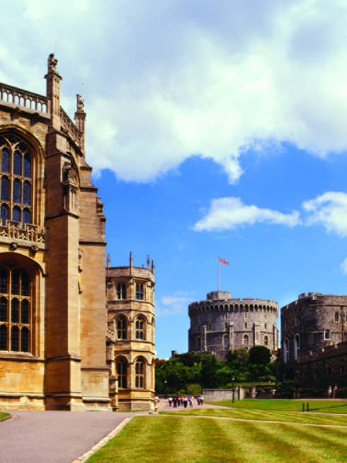 A wide shot of the Windsor Castle, with a manicured lawn, clear sky with large white coulds and a flag blowing in the background