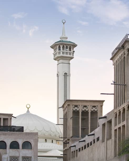 Street view of a mosque with a domed roof and adjoining minaret; in the fore, adobe buildings featuring ornate wind towers.