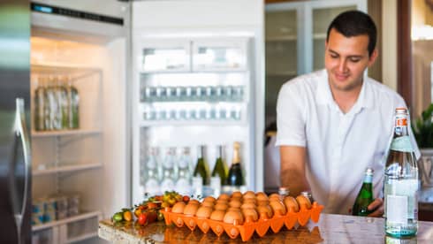 Smiling man holds green bottle in front of open fridge, fresh eggs on counter