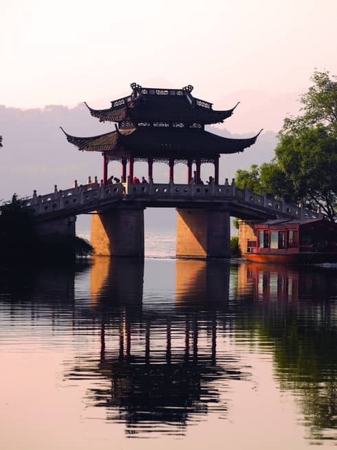 A boat floats towards a stone bridge with a pagoda cover cast in orange light from the setting sun.