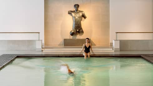 Indoor pool with a swimmer and a person sitting at the edge, featuring a large bronze torso sculpture on the far wall.