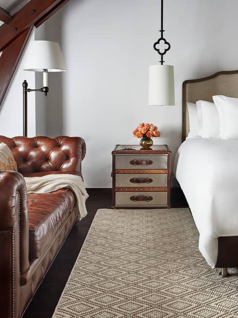 Brown period-style leather sofa under sloped beam roof next to nightstand, bed, cushioned bench