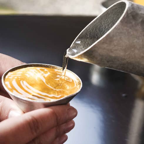 Clear liquid taking on amber colour as it is poured from a hammered metal spout into small cone-shaped cup held by two hands
