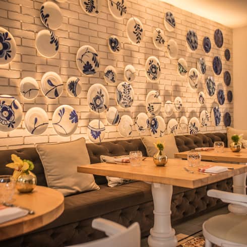 Restaurant dining room with white subray tile wall filled with blue-and-white painted plates, a long grey tufted bench and light-wood tables with white chairs on the other side of the bench