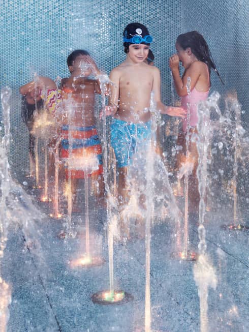 Six children play in a blue-tiled splash pad as water rises up from different holes in the floor