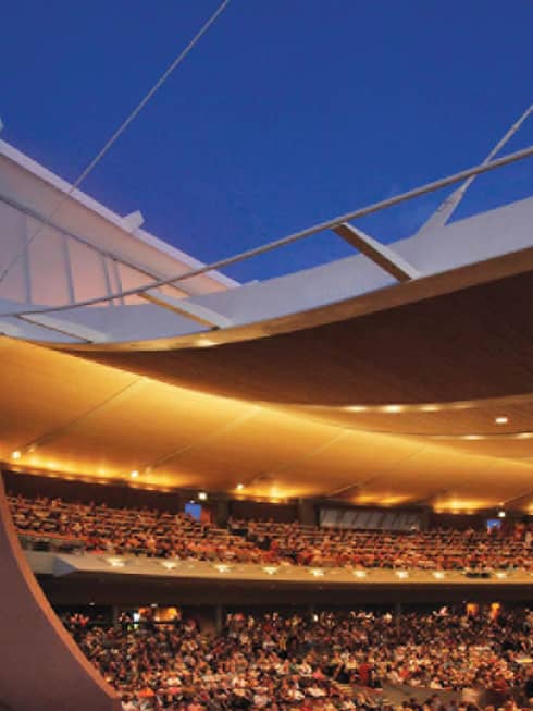 Curved opera building, audience in stands under lights at dusk