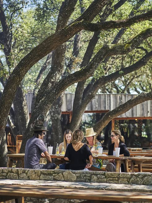 Guests sit and enjoy a meal on a wooden bench underneath an oak tree