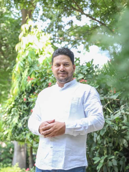 Person in long-sleeved white button-down stands with hands together outdoors in front of tree with red blooms