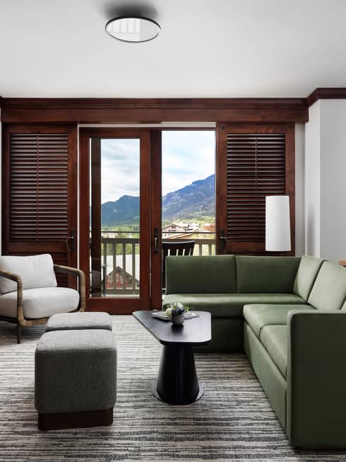 Bright sitting area with green sofa, armchair, two ottomans, coffee table, stone fireplace, TV and a balcony with mountain views through wooden shutters