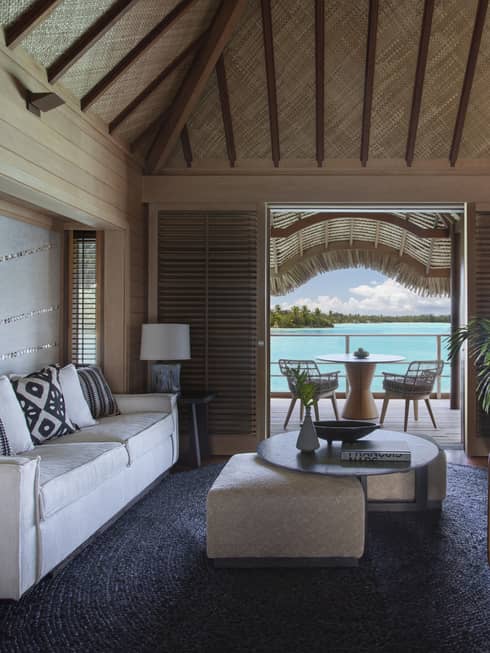 Overwater bungalow living area with neutral furniture, wood beams, tropical plants and a patio with a view of the turquoise lagoon.