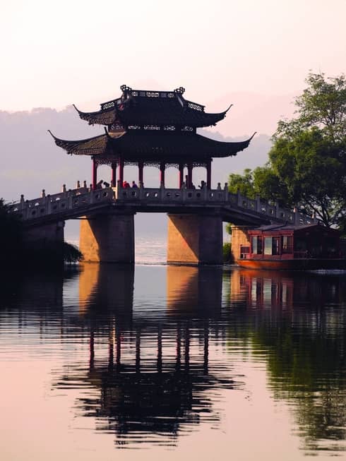 A boat floats towards a stone bridge with a pagoda cover cast in orange light from the setting sun.