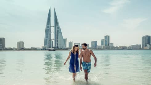 A man and woman run arm in arm through the shallow ocean water with Bahrain Bay skyscrapers in the background