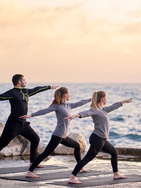 Three people in warrior yoga pose and coach standing at sea's edge at sunrise
