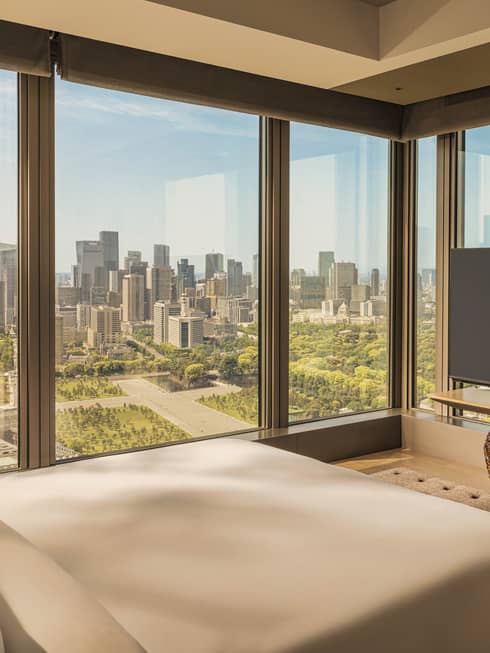 Luxury hotel room featuring king bed, TV and two walls of windows looking out to city and gardens